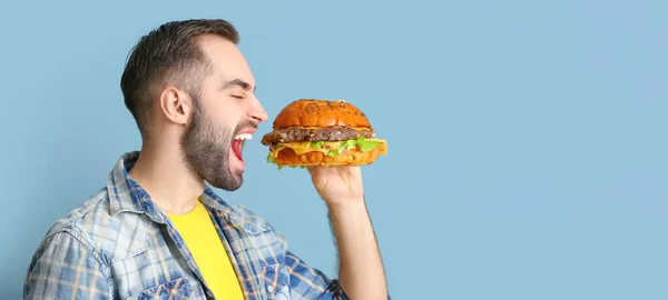 Young man eating tasty burger on light blue background with space for text