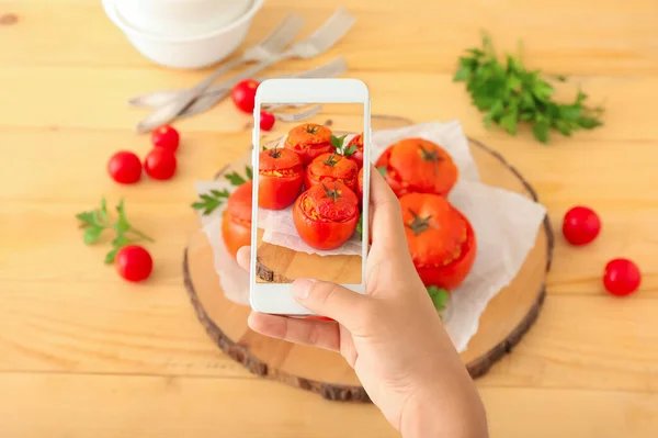 Female food photographer with mobile phone taking picture of tasty stuffed tomatoes on wooden table