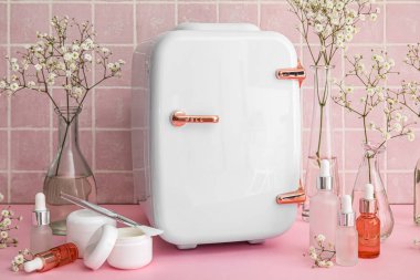 Doğal kozmetik ürünleri olan küçük bir buzdolabı ve pembe fayans duvarının yanındaki masada vazolarda çiçekler.