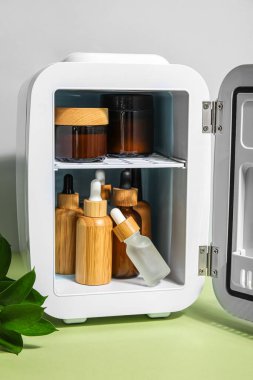 Gri duvarın yanındaki masada doğal kozmetik ürünleri ve bitki dalı olan küçük bir buzdolabı.