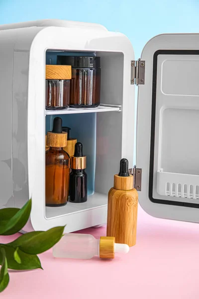 Small refrigerator with natural cosmetics on pink table near blue wall