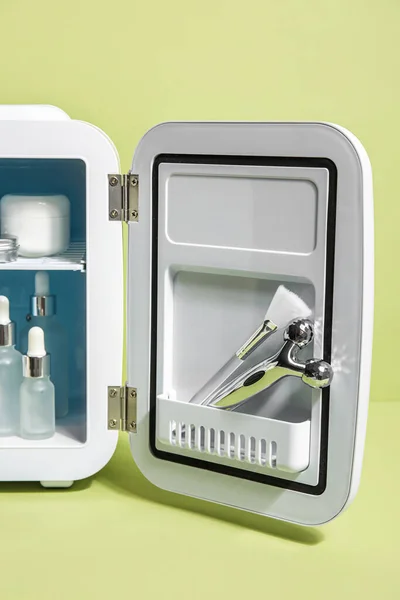 Small refrigerator with natural cosmetics, brush and massage tool on green background