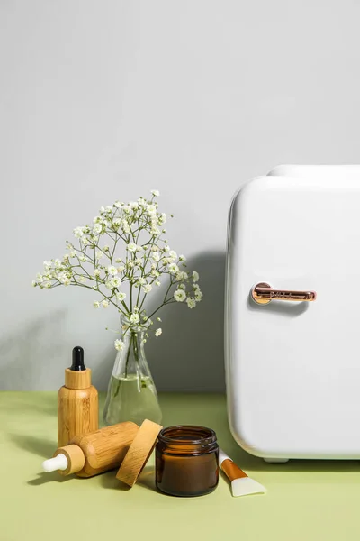 Small refrigerator with natural cosmetics and flowers in vase on table near grey wall
