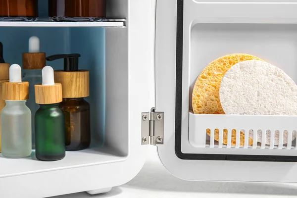 Small refrigerator with natural cosmetics and sponges on white background, closeup