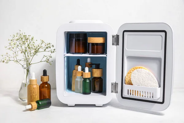 Small refrigerator with natural cosmetics and flowers in vase on white background