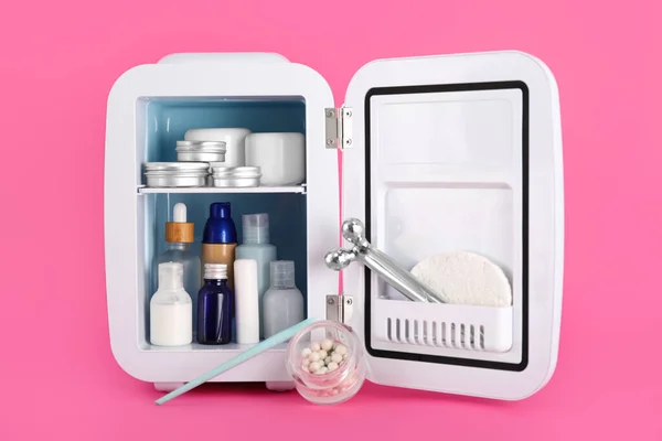 Small refrigerator with cosmetic products on pink background