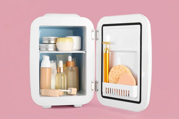 Small refrigerator with cosmetic products on pink background