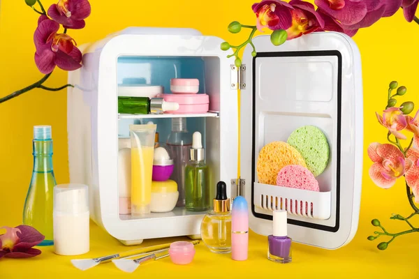 Small refrigerator with cosmetics and flowers on yellow background