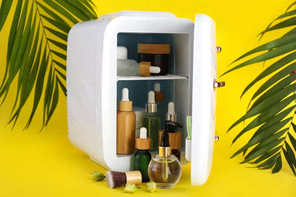 Small refrigerator with cosmetics and tropical palm leaves on yellow background