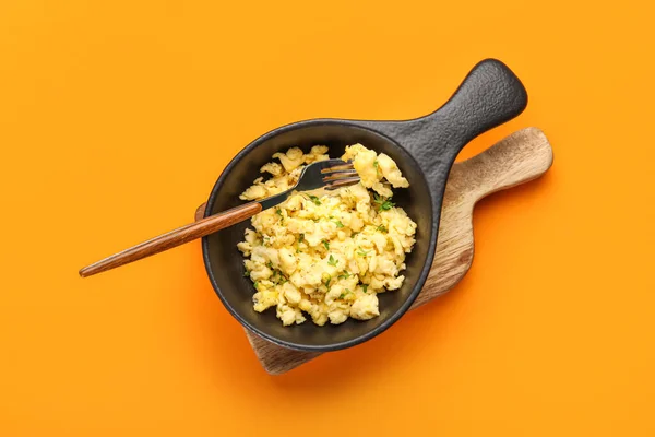 Wooden board with frying pan of tasty scrambled eggs on color background