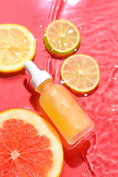 Bottle of vitamin C serum and fruits in water on red background, closeup