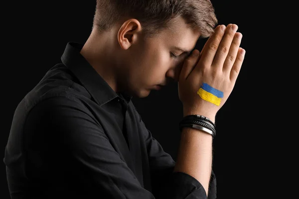Young man with drawn flag of Ukraine praying on black background, closeup