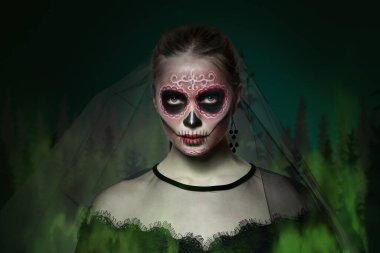 Meksika Ölüler Günü 'nde yüzünde boyalı kafatası olan genç bir kadın koyu yeşil arka plana karşı.