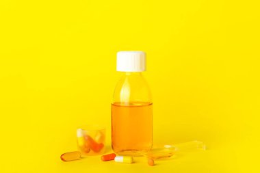 Bir şişe öksürük şurubu, fincan, kaşık ve sarı arka planda haplar.