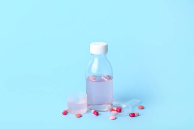 Bir şişe öksürük şurubu, fincan, kaşık ve mavi arka planda haplar.