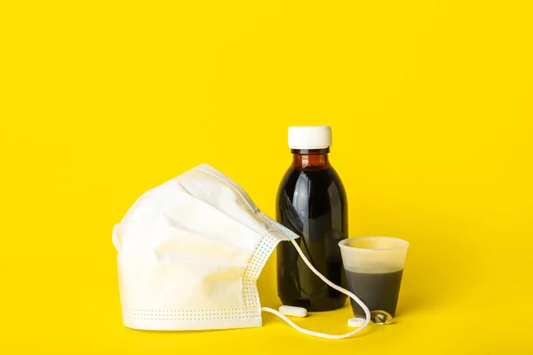 Bottle of cough syrup, cup, pills and medical mask on yellow background
