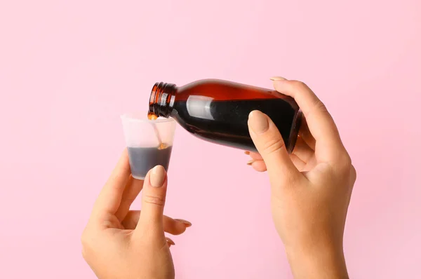 Woman pouring cough syrup from bottle into cup on pink background