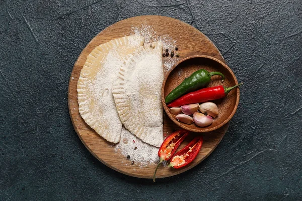 Board with raw chebureks, bowl of chili peppers and garlic on dark background