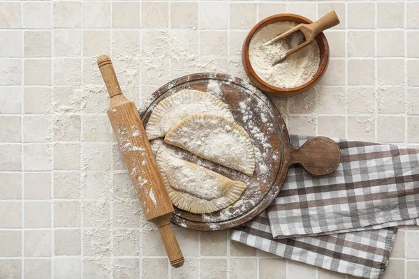 Board with raw chebureks, rolling pin and bowl of flour on beige tile background