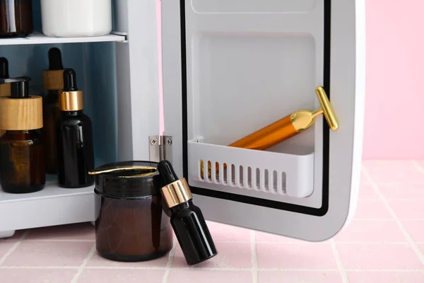 Small refrigerator with natural cosmetics and facial massage tool on pink tile table, closeup