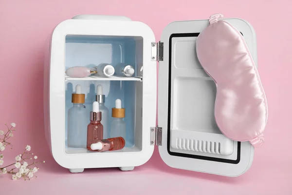 Small refrigerator with cosmetic bottles, facial massage tool and sleeping mask on pink background