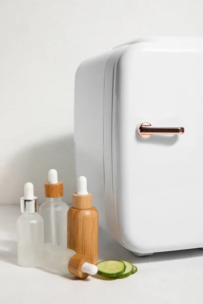 Small refrigerator with cosmetic dropper bottles and cucumber slices on white background