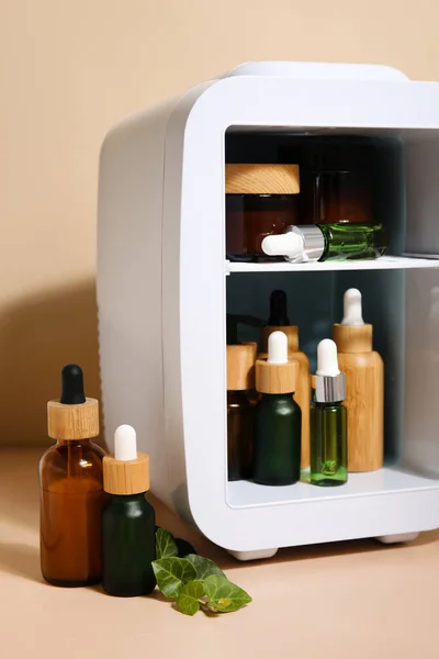 Small refrigerator with natural cosmetics and plant leaves on beige background