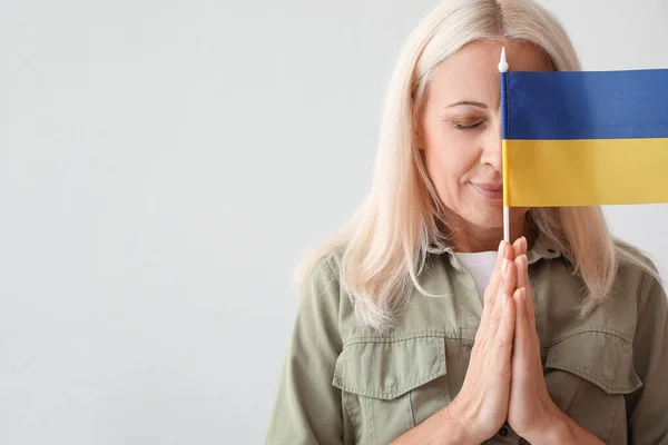 Mature woman with flag of Ukraine praying on light background