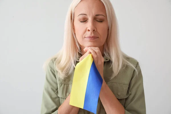 Mature woman with flag of Ukraine praying on light background