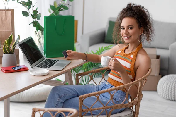 Young African-American woman with cup of coffee and credit card shopping online at home