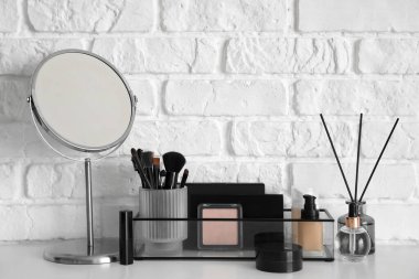 Makyaj fırçaları, kozmetik ürünleri, ayna ve kamış ayırıcılı tutucu beyaz tuğla duvarın yanındaki masada.