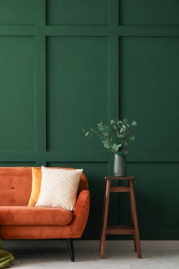 Masada okaliptüs dalları ve yeşil duvarın yanında kırmızı kanepe olan vazo.