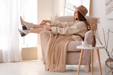 Zarif giysiler içinde güzel, modaya uygun genç bir kadın evdeki koltukta oturuyor.
