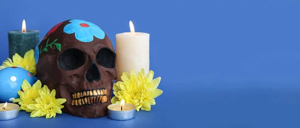 Painted skull for Mexico\'s Day of the Dead (El Dia de Muertos), flowers and burning candles on blue background with space for text