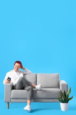 Sıra dışı saçları ve telefonu olan güzel genç bir kadın mavi arka planda koltukta oturuyor.