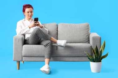Sıra dışı saçları ve telefonu olan güzel genç bir kadın mavi arka planda koltukta oturuyor.
