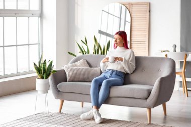 Alışılmadık saçları olan güzel genç bir kadın evdeki kanepede dinlenirken kahve içiyor.