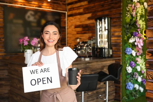 Beautiful business owner with opening sign and coffee cup in her cafe