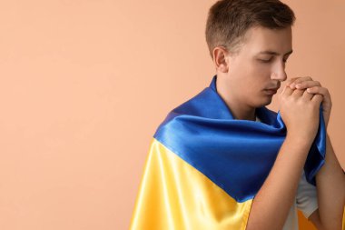Ukrayna bayrağı taşıyan genç adam bej arka planda dua ediyor.