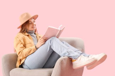 Pembe arka planda koltukta kitap okuyan genç bir kadın.