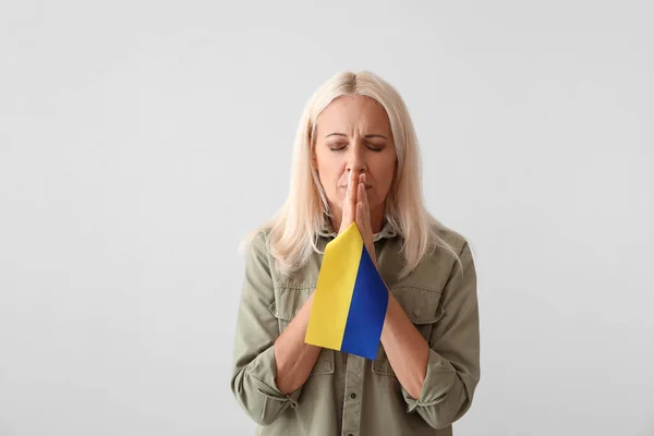 Mature woman with flag of Ukraine praying on light background