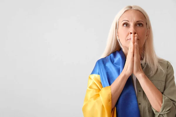 Mature woman with flag of Ukraine praying on light background