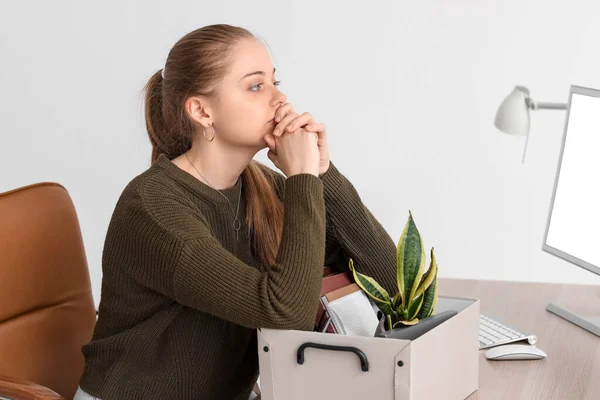 Upset fired young woman with packed stuff sitting at table in office