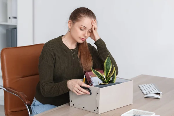 Upset fired young woman with packed stuff sitting at table in office