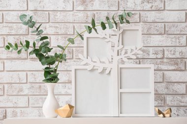 Fotoğraf çerçeveli aile ağacı, vazoda okaliptüs dalları ve tuğla duvarın yanındaki masada dekor.