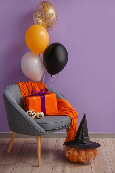 Armchair with pumpkins, gift and Halloween balloons near violet wall