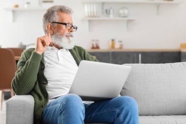 Bilgisayarlı, düşünceli, olgun bir adam mutfaktaki kanepede oturuyor.