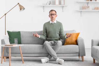 Genç adam evdeki kanepede meditasyon yapıyor.