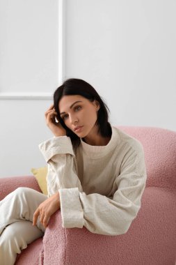 Bej koltukta oturan genç bir kadın aydınlık duvarın yanında