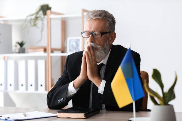 Зрелый человек с флагом Украины молится в своем кабинете